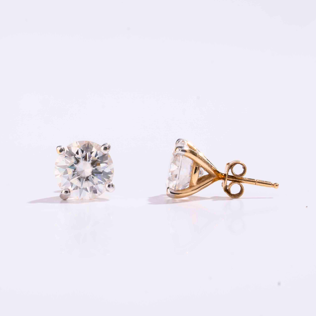 A pair of round solitaire four prong martini setting earrings with a gold metal band and a clear, sparkling gemstone in a martini setting.